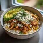 A close-up of a street corn chicken rice bowl topped with shredded chicken, crumbled feta, cilantro, and sliced avocado.