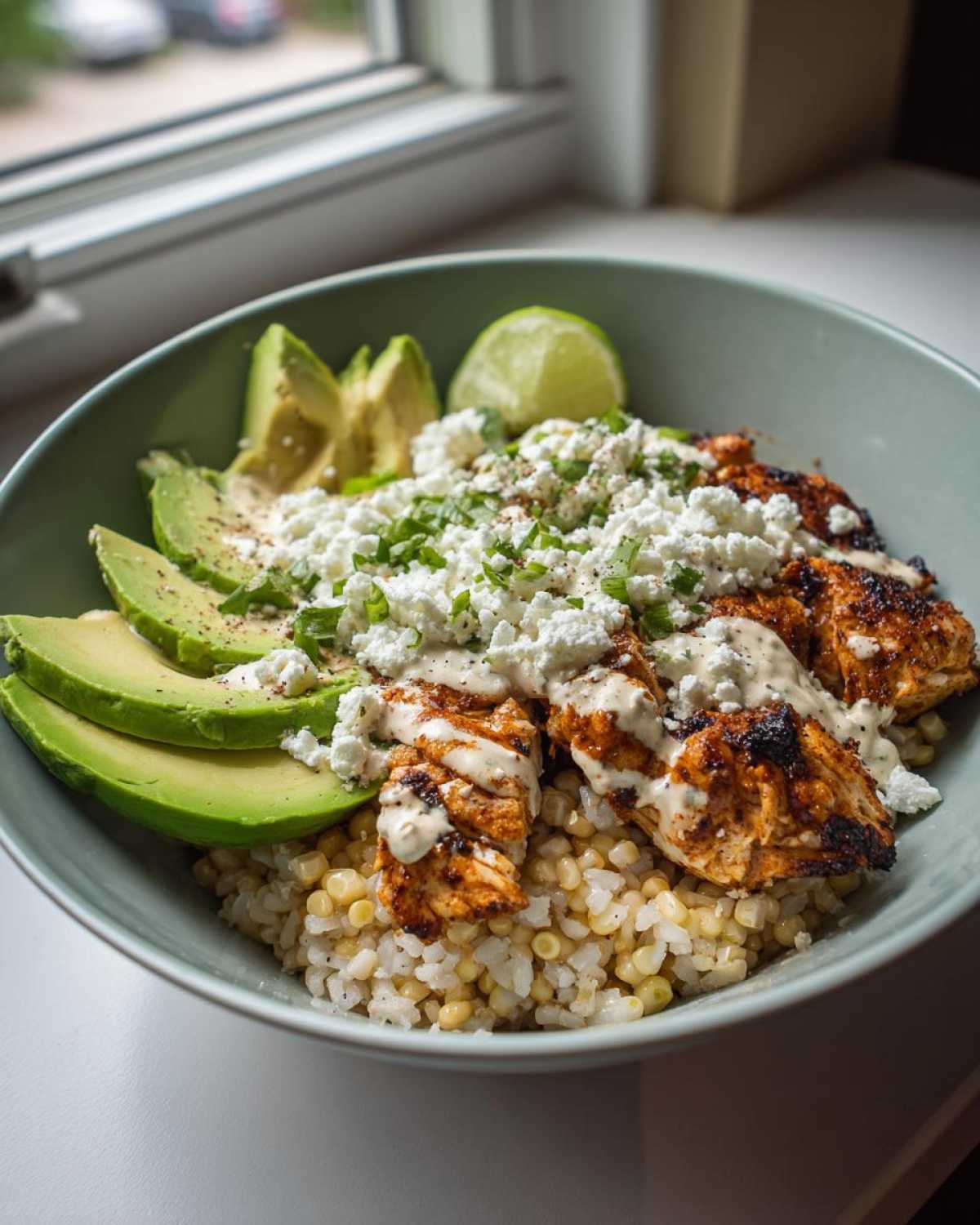 A vibrant street corn chicken rice bowl featuring grilled chicken, corn, avocado slices, crumbled feta, and a drizzle of sauce.