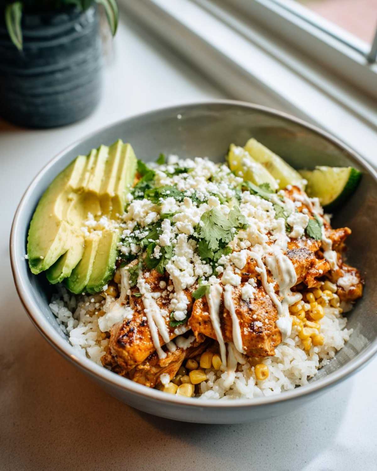 A delicious street corn chicken rice bowl topped with avocado slices, crumbled cheese, cilantro, and lime wedges.