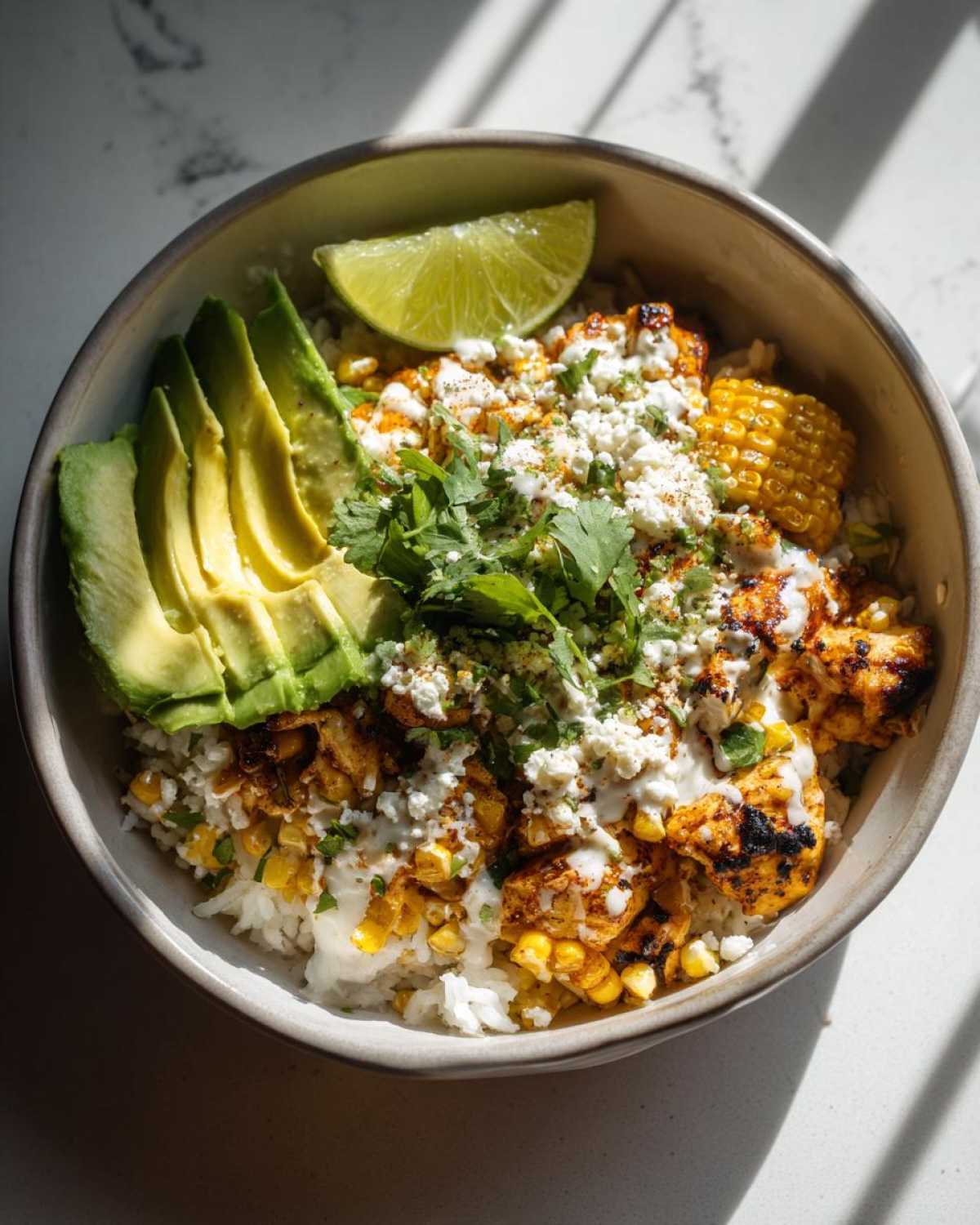 A vibrant street corn chicken rice bowl topped with sliced avocado, crumbled cheese, cilantro, and a lime wedge.