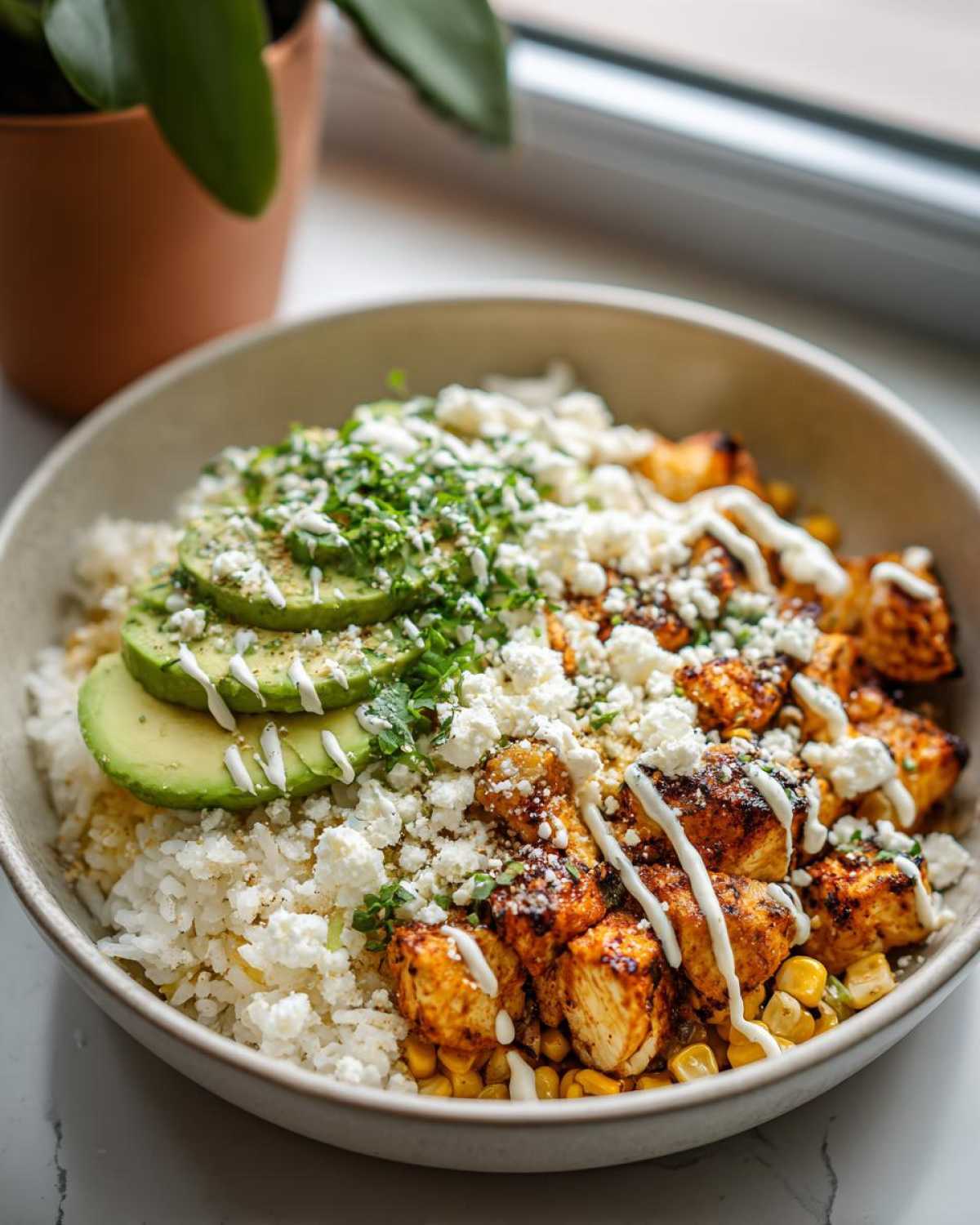 A delicious Street Corn Chicken Rice Bowl featuring seasoned chicken, white rice, corn, avocado slices, crumbled feta, and a drizzle of crema.