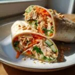 A close-up of two halves of a Thai peanut chicken wrap, filled with shredded chicken, carrots, cucumber, and peanuts.