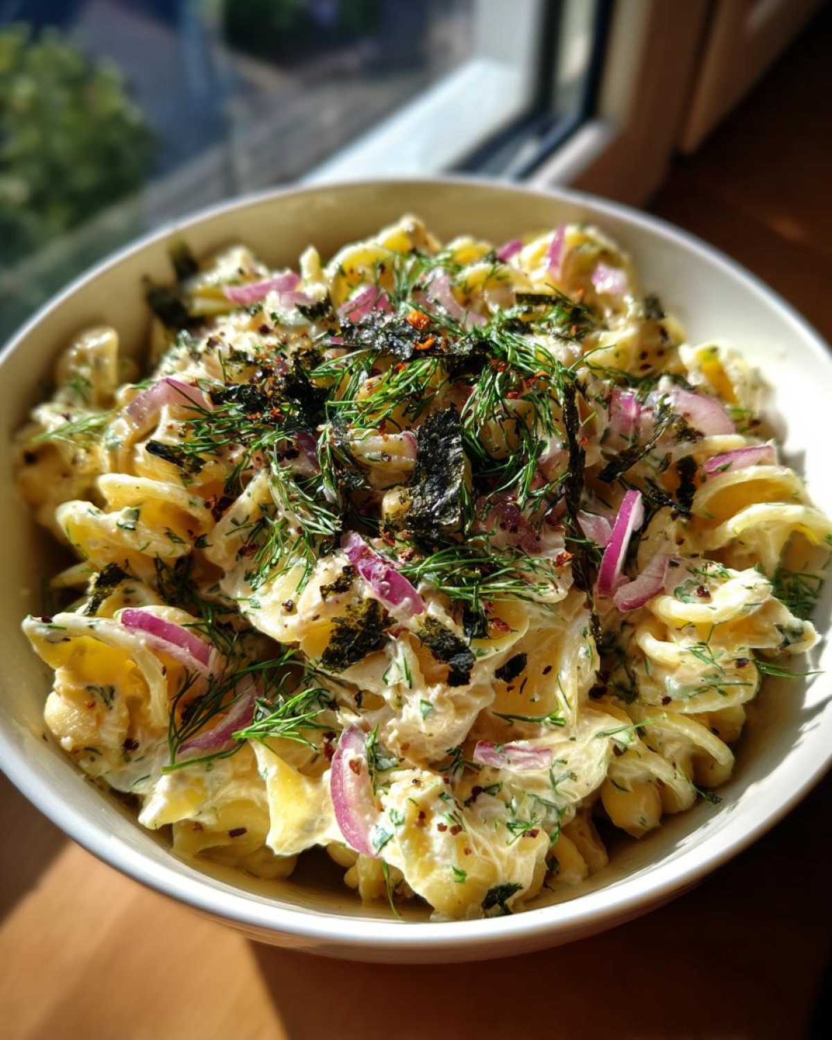 A close-up of a bowl filled with creamy vegan tuna pasta salad, featuring fusilli pasta, red onion, dill, and seaweed flakes.