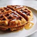 Close-up of a golden-brown vegan waffle stacked on a white plate, generously drizzled with syrup.