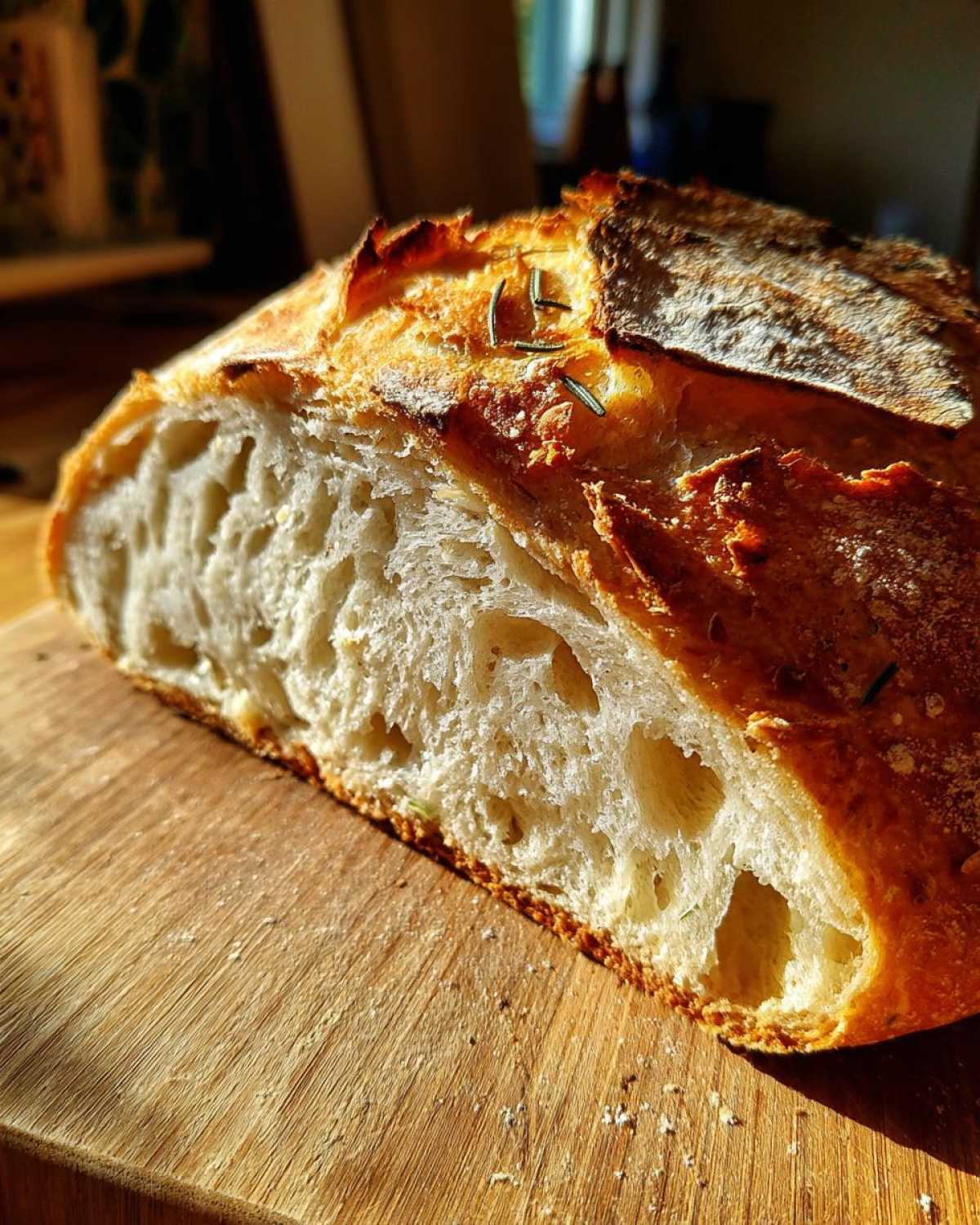 Close-up of a sliced whole wheat rosemary bread with a golden crust and airy crumb on a wooden board.