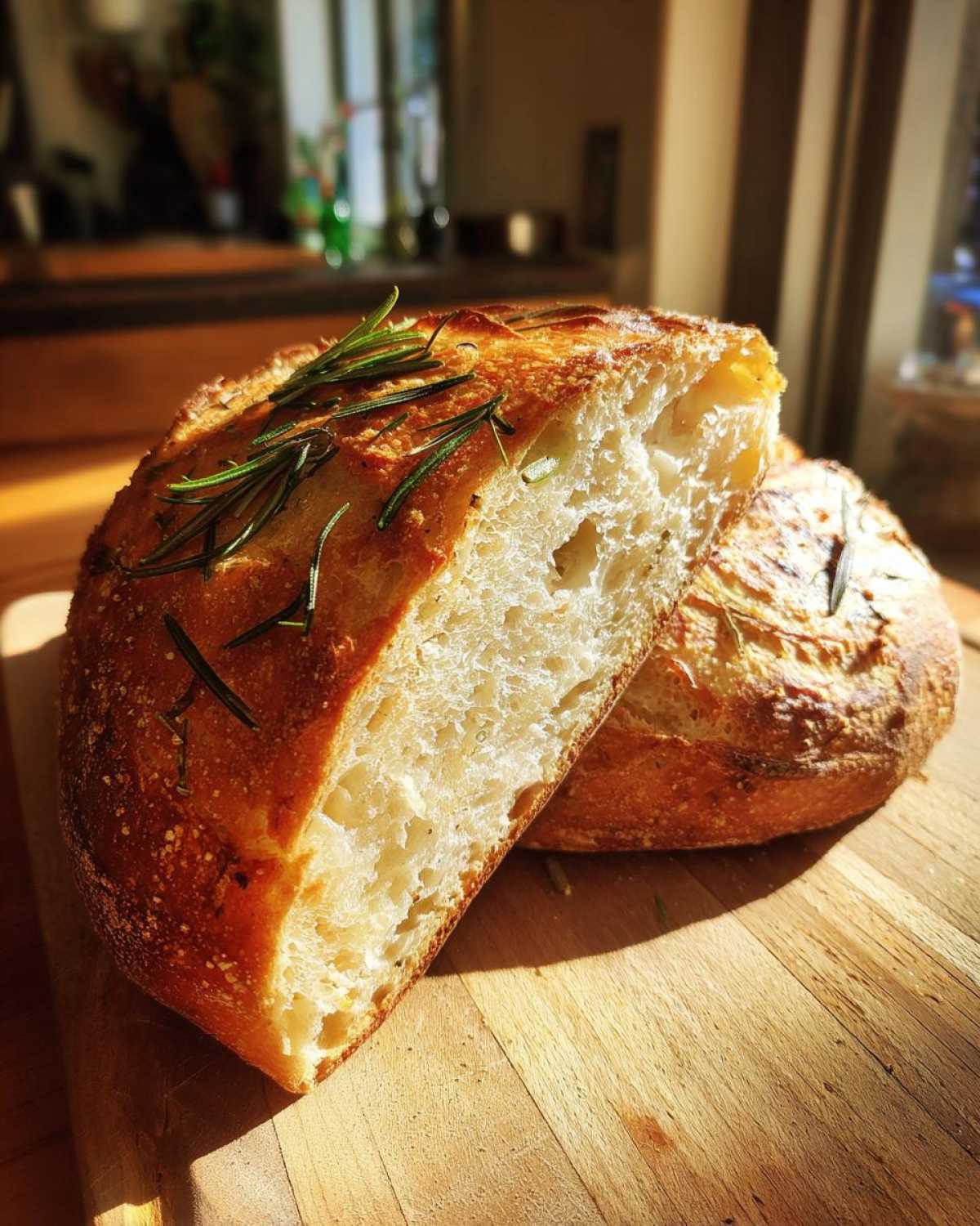 A close-up of a freshly baked whole wheat rosemary bread, cut in half, showing its airy crumb and golden crust.