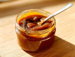 A close-up of a glass jar filled with rich, dark apple butter, with a spoon resting inside.