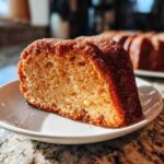 A close-up of a slice of moist Apple Cider Donut Cake, coated in cinnamon sugar, on a white plate.