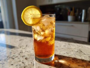 A tall glass filled with ice and amber-colored Apple Cider Long Island Iced Tea, garnished with a lemon slice.
