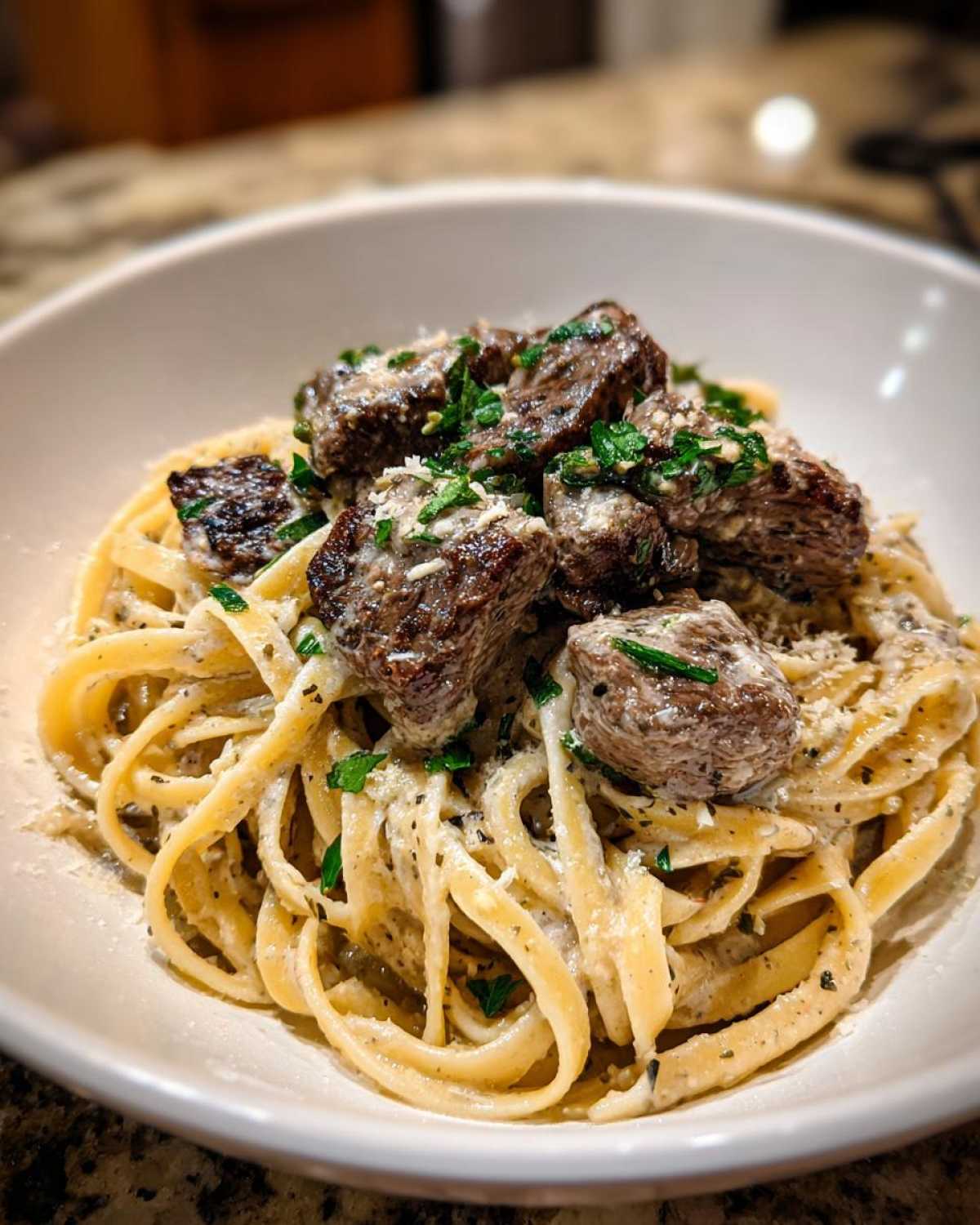 A close-up of a white bowl filled with beef garlic butter linguine, topped with tender beef chunks and fresh parsley.