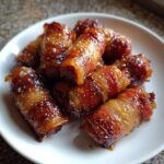 A close-up of a pile of glistening brown sugar bacon little smokies on a white plate.