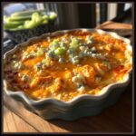 A close-up of a bubbling classic Buffalo chicken dip in a baking dish, topped with melted cheese, blue cheese crumbles, and chopped celery.