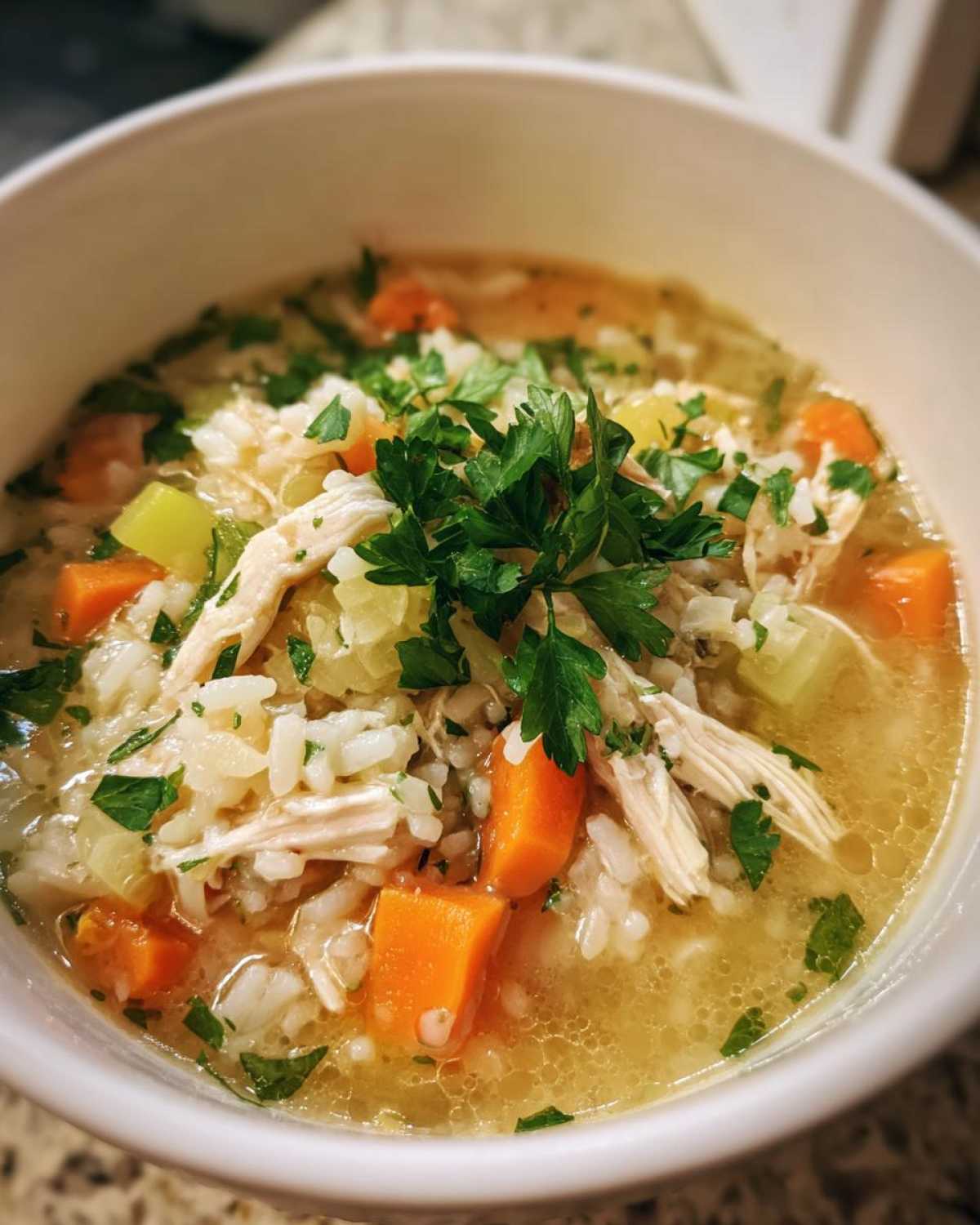 A close-up of a bowl of hearty chicken and rice soup, filled with shredded chicken, rice, carrots, celery, and fresh parsley.