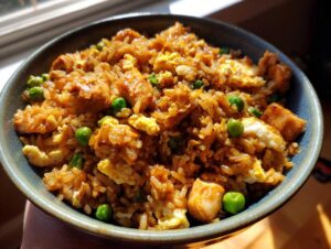 A close-up shot of a bowl filled with savory chicken fried rice, featuring tender chicken pieces, scrambled eggs, and bright green peas.