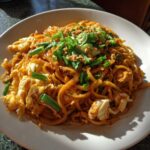 A close-up of a plate of Chicken Pad Thai, featuring noodles, scrambled egg, and green onions.