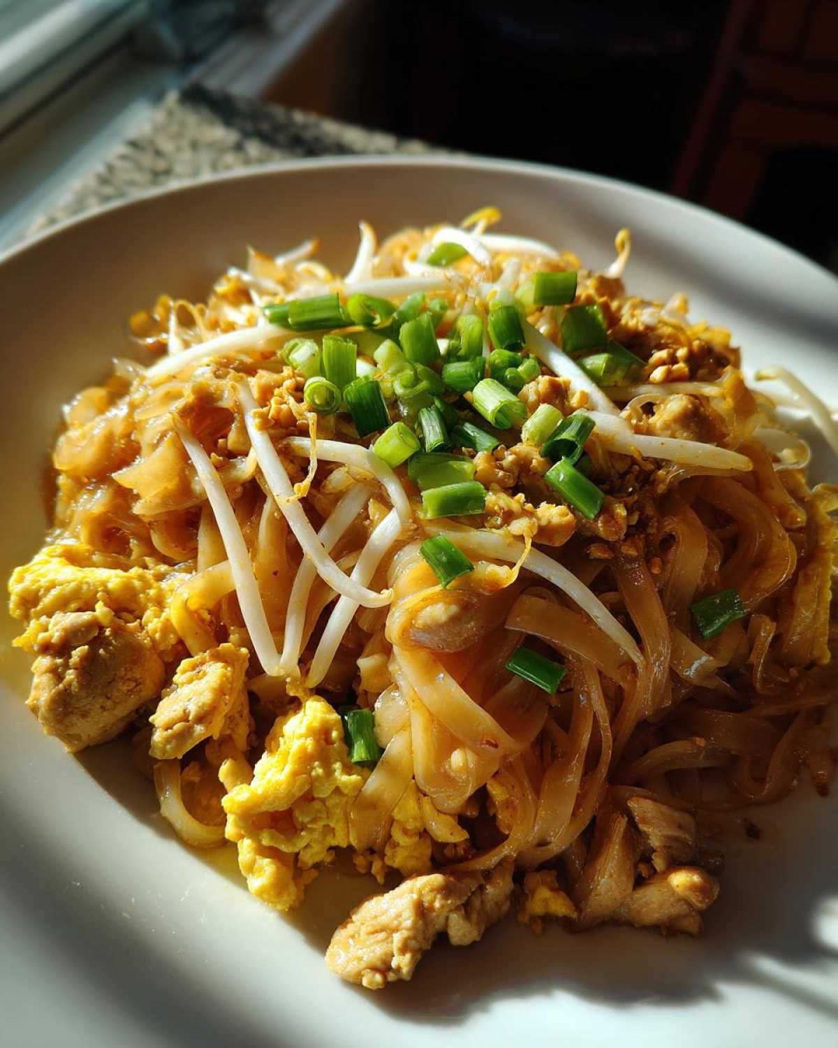 Close-up of a plate of Chicken Pad Thai, featuring rice noodles, scrambled egg, chicken pieces, bean sprouts, and chopped green onions.