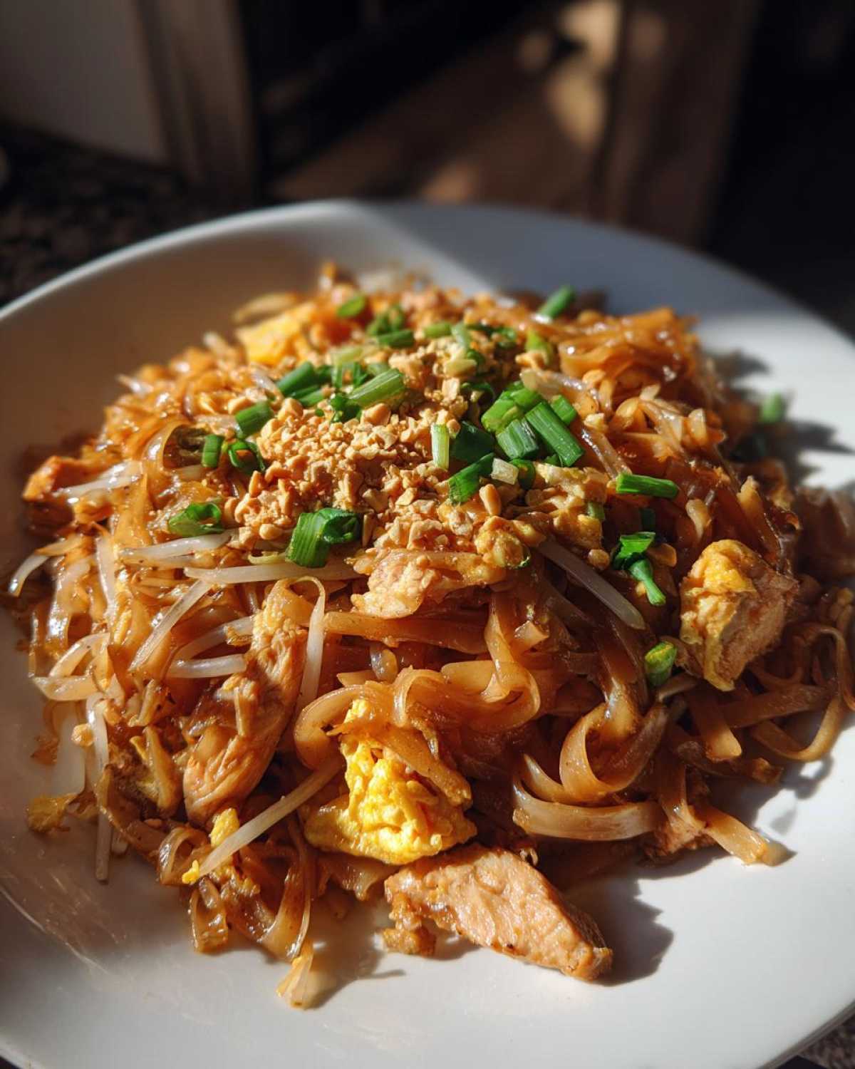 A close-up of a serving of Chicken Pad Thai, featuring noodles, chicken, scrambled egg, bean sprouts, crushed peanuts, and chopped scallions.