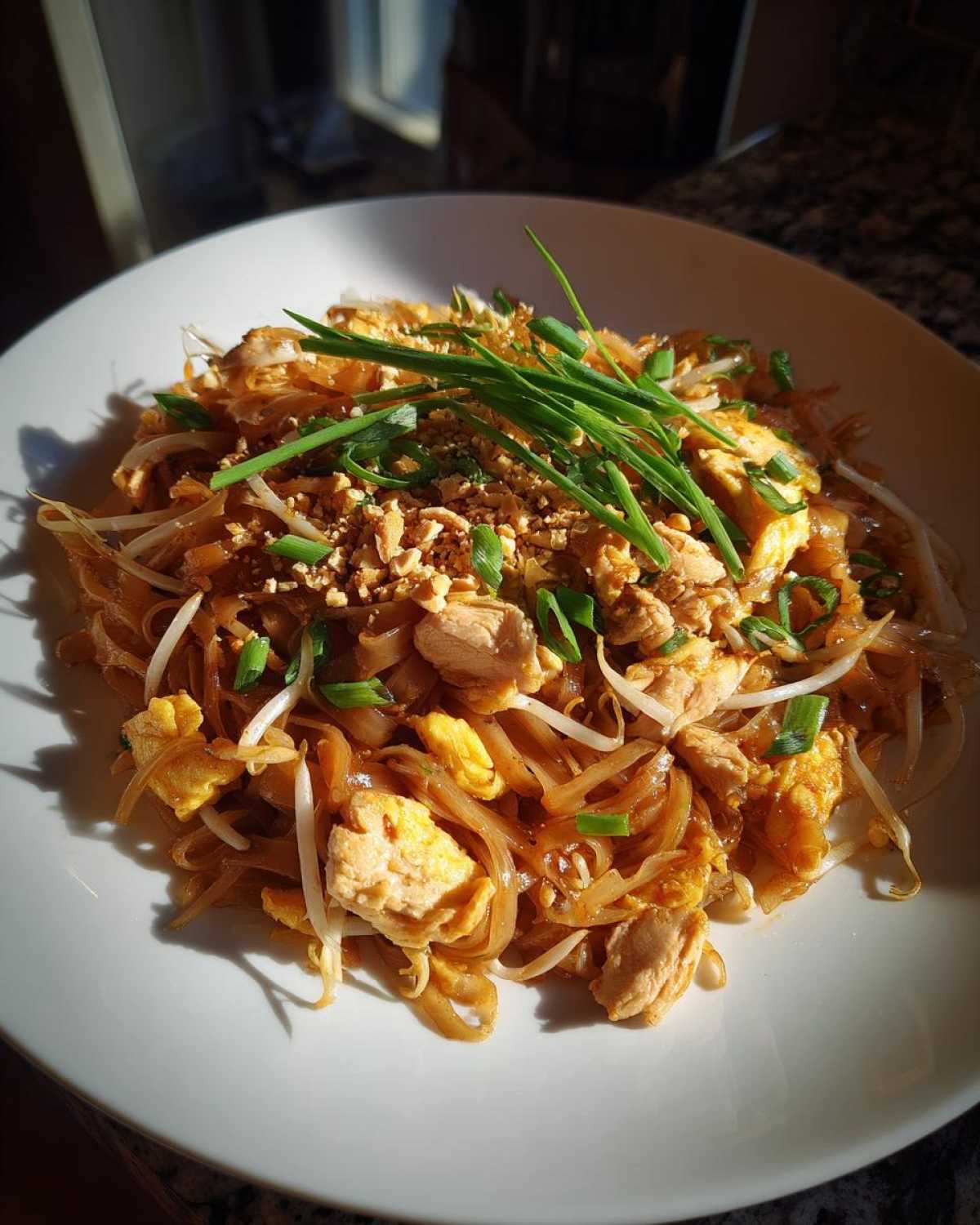 A close-up of a white plate filled with Chicken Pad Thai, featuring noodles, chicken, bean sprouts, and green onions.
