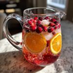 A clear glass pitcher filled with sparkling cranberry orange punch, ice, fresh cranberries, and orange slices.