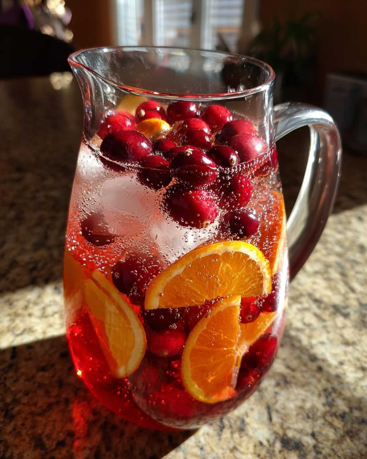 A clear glass pitcher filled with bubbly Cranberry Orange Punch, ice, fresh cranberries, and orange slices.