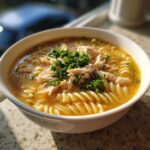 A close-up of a bowl of creamy chicken noodle soup, topped with shredded chicken and fresh parsley.