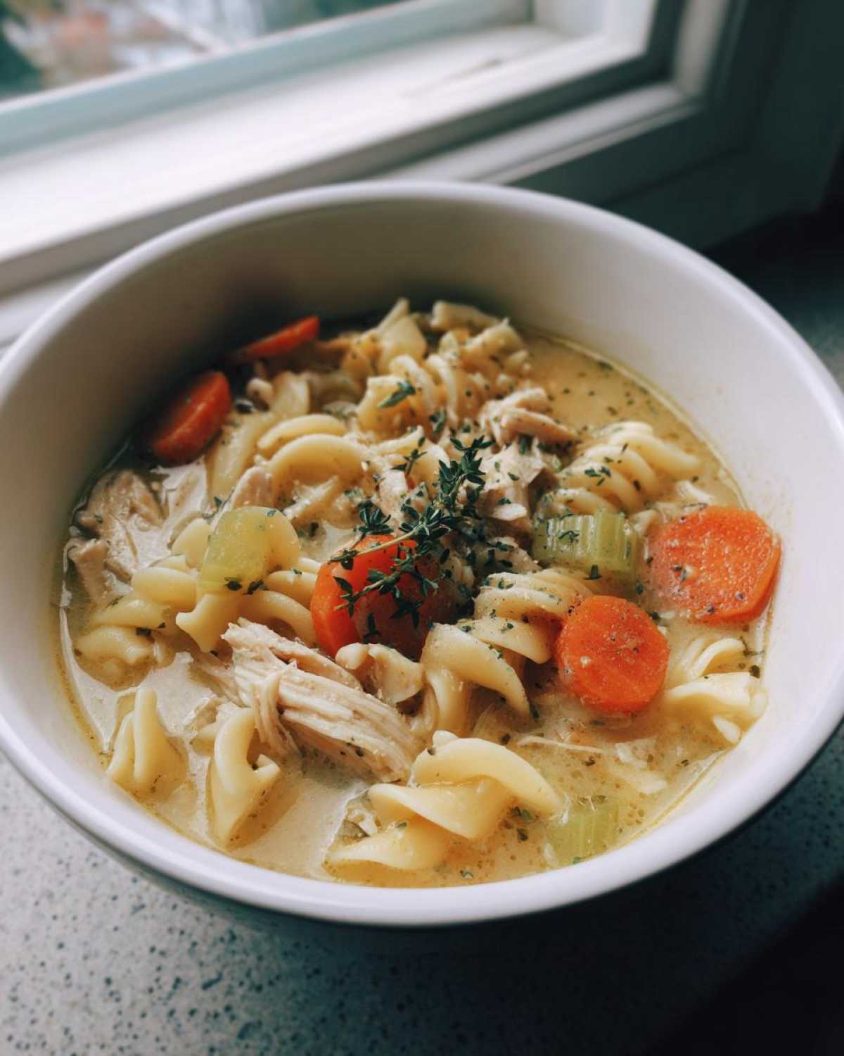 A close-up of a bowl of creamy chicken pasta soup with rotini noodles, shredded chicken, carrots, and celery, garnished with herbs.