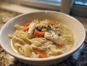 A close-up of a white bowl filled with creamy chicken pasta soup, featuring noodles, shredded chicken, carrots, and celery, garnished with thyme.