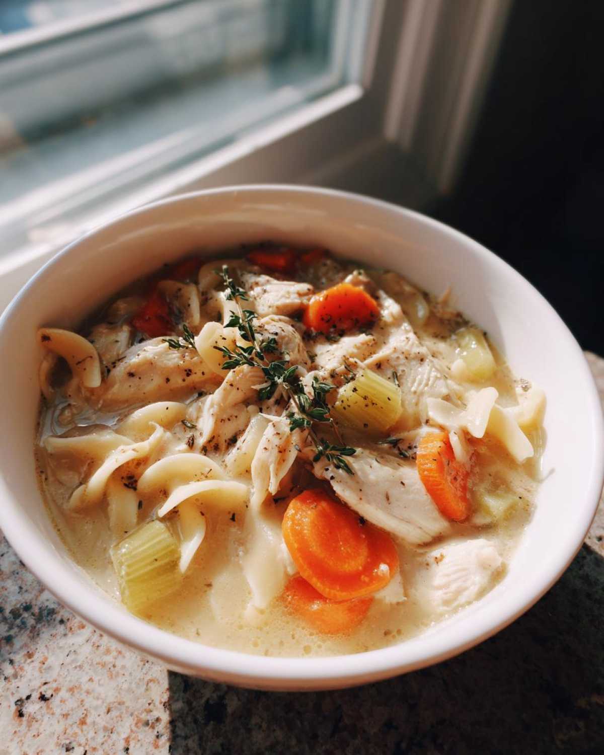 A close-up of a bowl of creamy chicken pasta soup with noodles, shredded chicken, carrots, and celery, garnished with thyme.