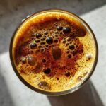 Close-up overhead view of a glass filled with creme brulee New Orleans iced coffee, showing rich brown liquid topped with a foamy crema and bubbles.