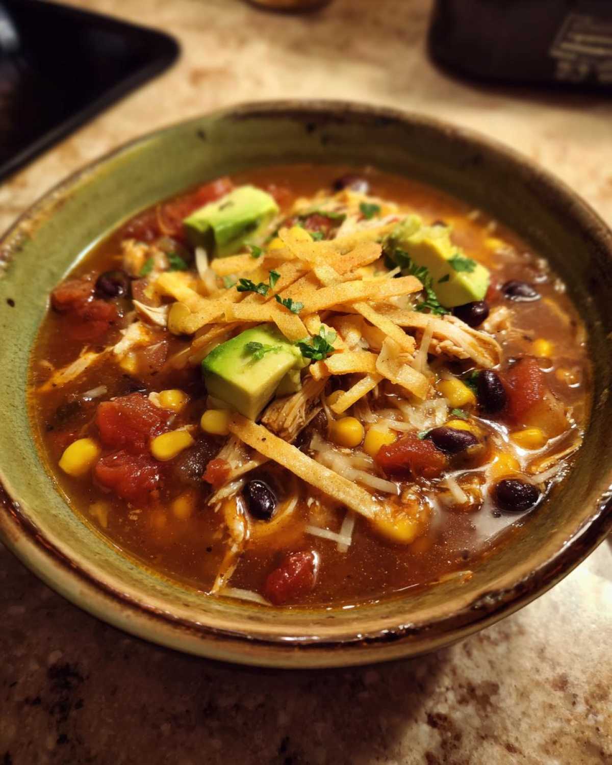 A close-up of a bowl of hearty crock pot chicken tortilla soup, topped with avocado, tortilla strips, cheese, and cilantro.