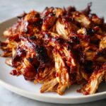 A close-up of shredded crockpot BBQ chicken, glistening with sauce on a white plate.