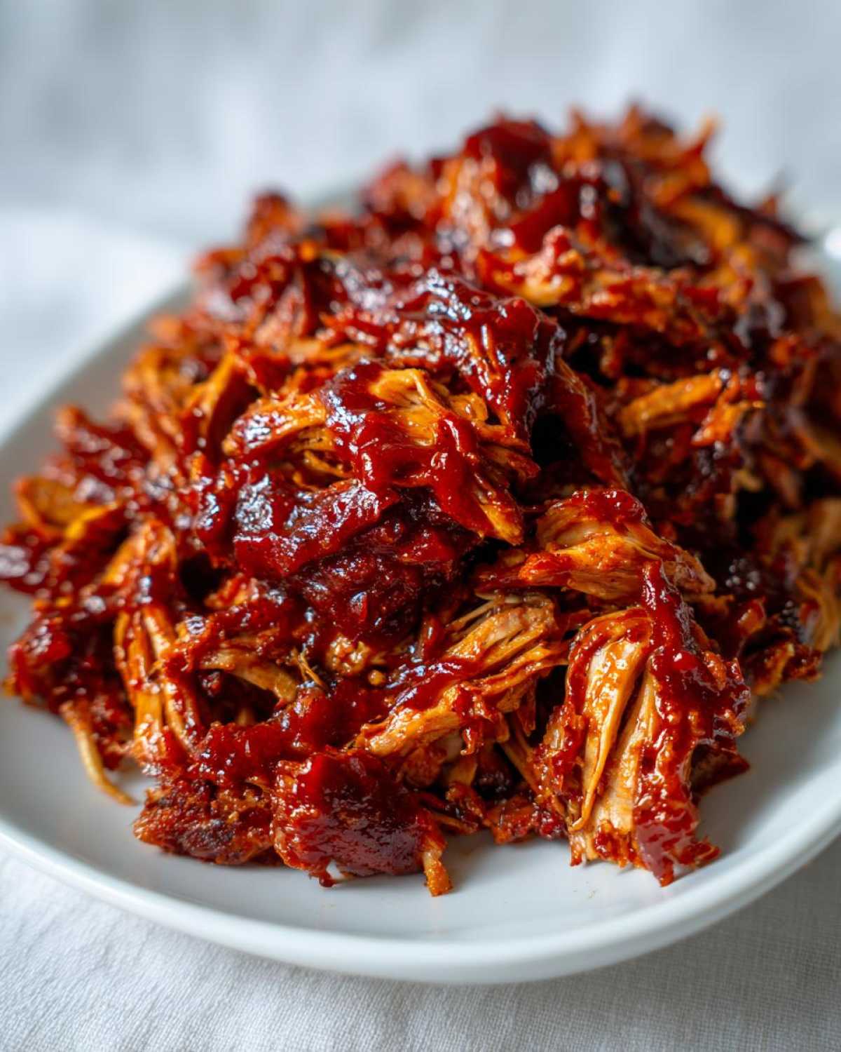A close-up of tender, shredded crockpot BBQ chicken coated in rich, glossy BBQ sauce on a white plate.