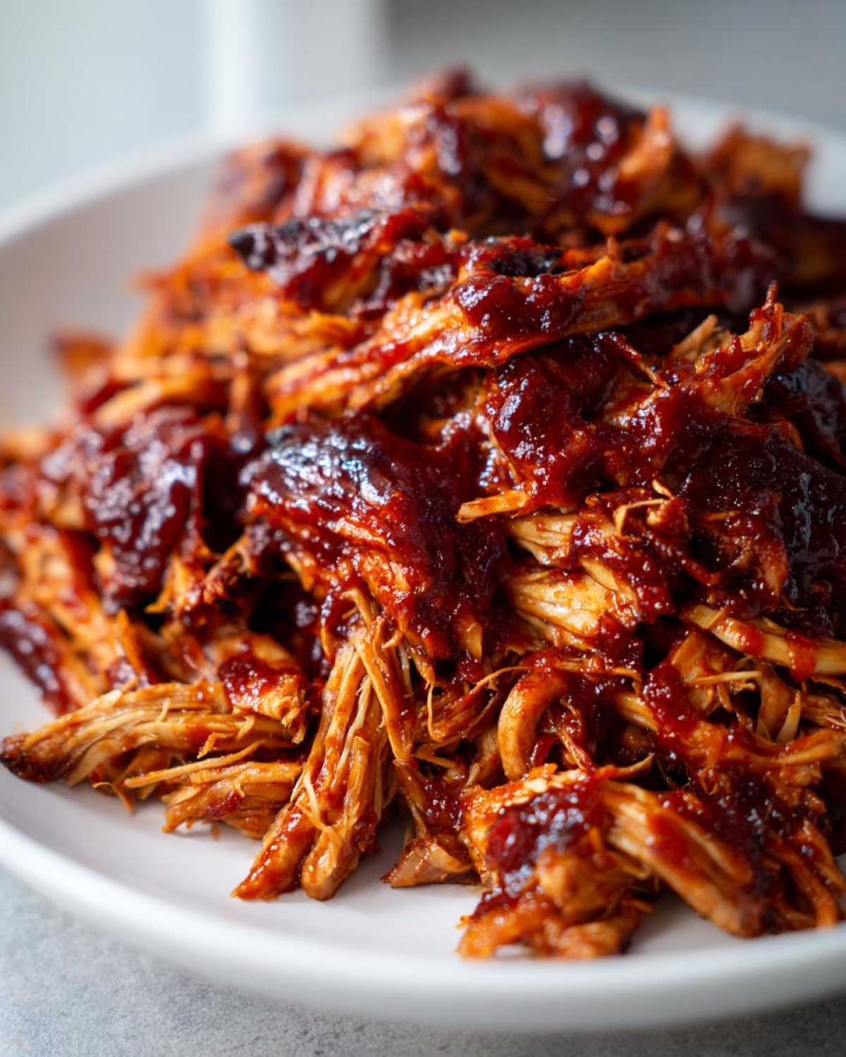 Close-up of shredded crockpot BBQ chicken coated in rich, glossy BBQ sauce on a white plate.