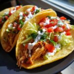 Three fully loaded crockpot chicken tacos with shredded chicken, lettuce, tomatoes, and sour cream on a black plate.