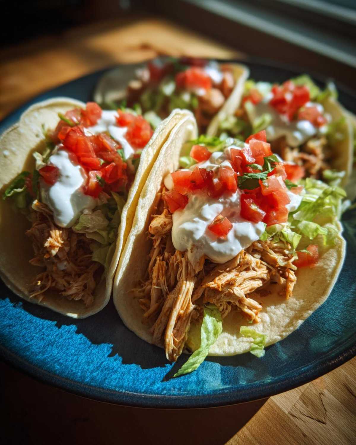 Close-up of three delicious crockpot chicken tacos filled with shredded chicken, lettuce, sour cream, and diced tomatoes.