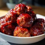 A close-up of Crockpot grape jelly meatballs glistening in a white bowl, coated in a rich, dark glaze.