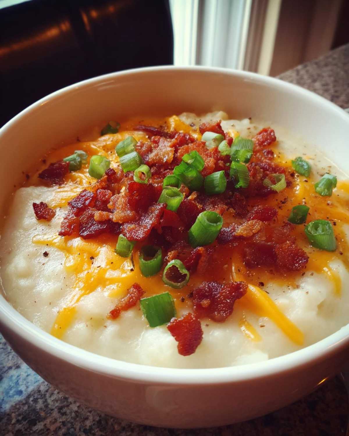A close-up of a bowl of creamy Crockpot potato soup, generously topped with shredded cheddar cheese, crispy bacon bits, and chopped green onions.