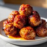 A close-up shot of a pile of glistening crockpot sweet chili meatballs on a white plate.