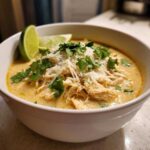 A close-up of a white bowl filled with Crockpot white chicken chili, topped with shredded cheese, cilantro, and lime wedges.