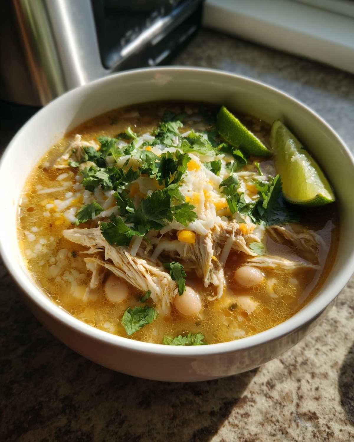 A close-up of a bowl of Crockpot white chicken chili, topped with shredded cheese, cilantro, and lime wedges.