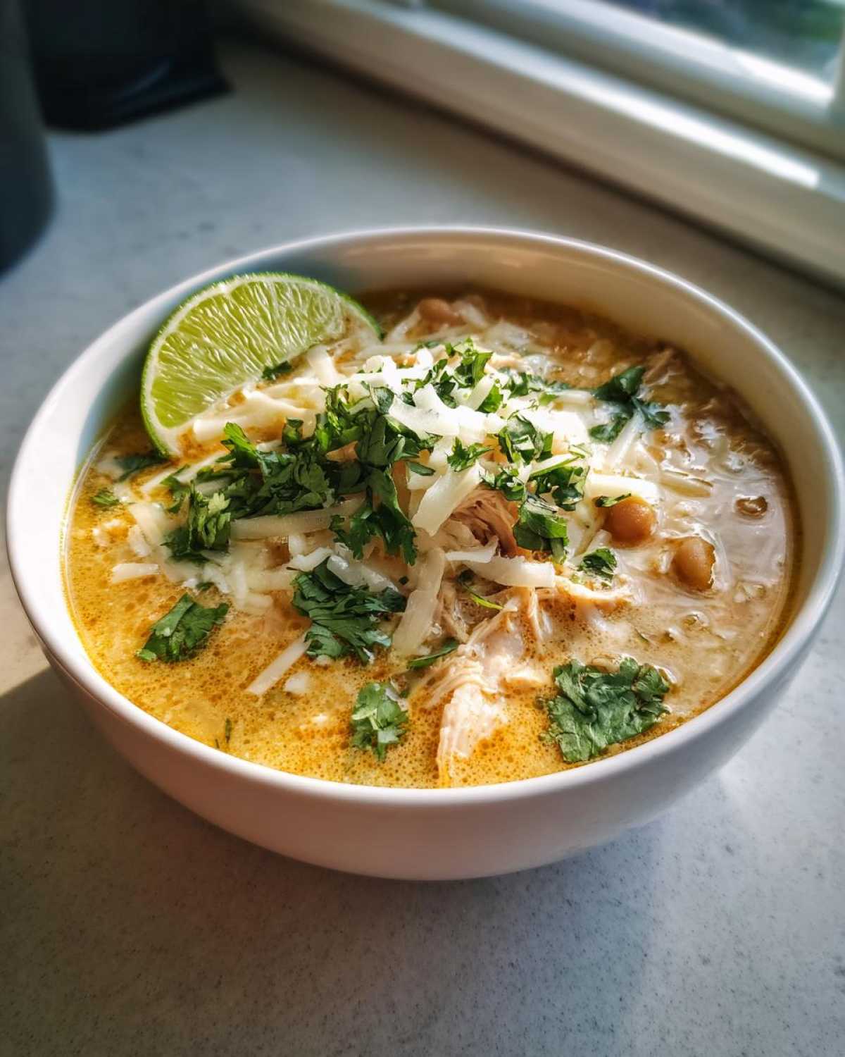 A bowl of Crockpot white chicken chili topped with shredded cheese, fresh cilantro, and a lime wedge.
