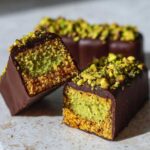 Close-up of a decadent Dubai chocolate bar cut in half, revealing pistachio cream filling and a crunchy base, topped with chopped pistachios.