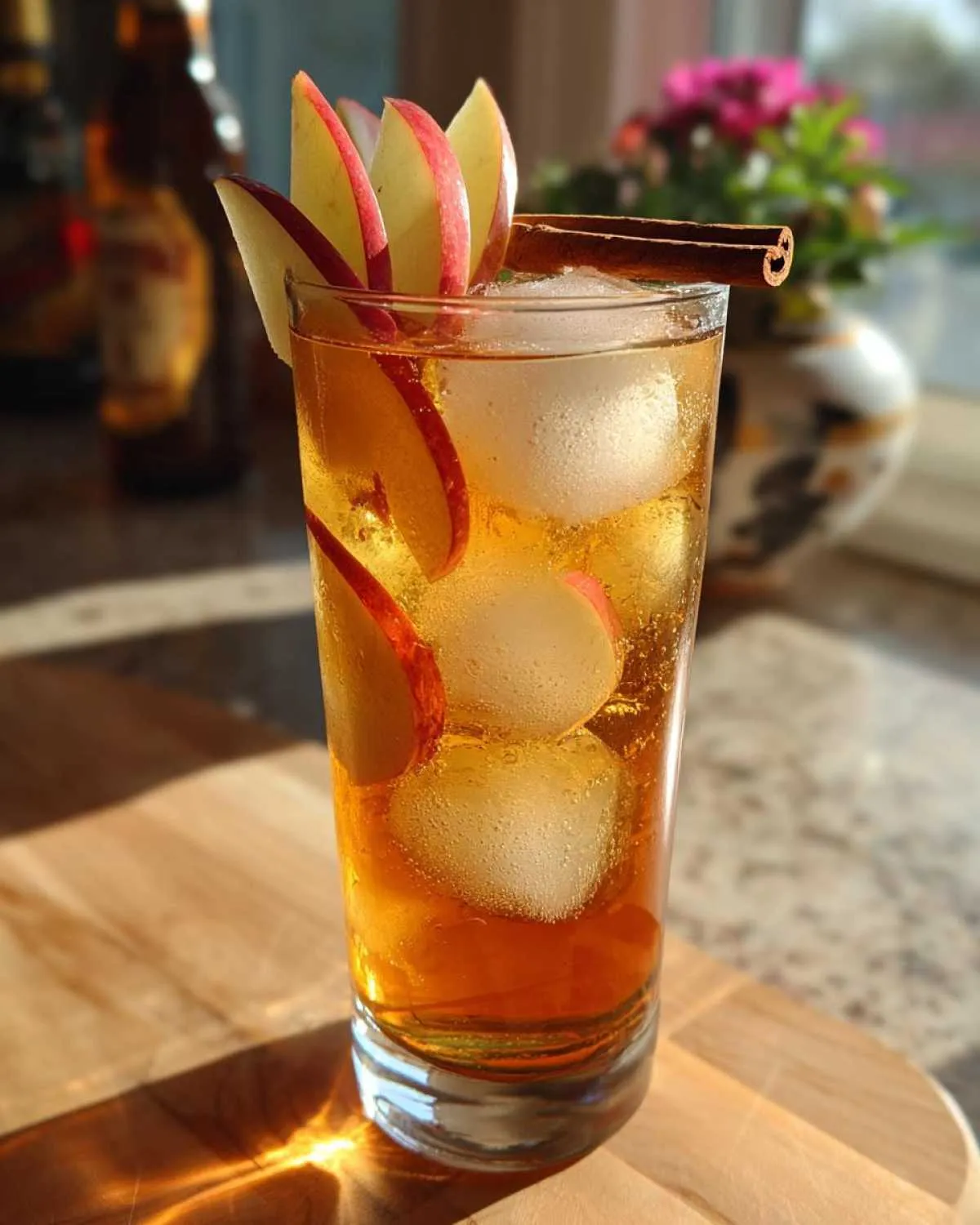 A tall glass of Fall Long Island Iced Tea garnished with apple slices and a cinnamon stick, filled with ice and amber liquid.