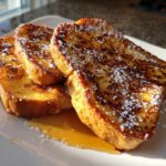 Three slices of golden brown French toast dusted with powdered sugar and drizzled with syrup.