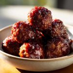 A close-up of a bowl filled with glistening grape jelly meatballs, coated in a rich, savory sauce.