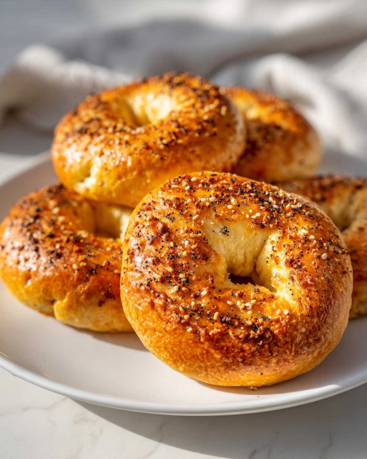 A close-up of several golden-brown Greek yogurt bagels topped with everything bagel seasoning on a white plate.