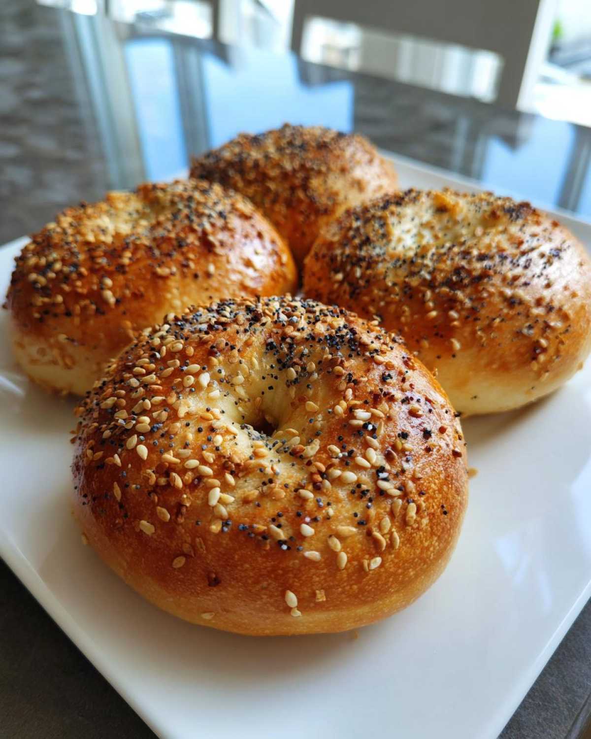 Four golden-brown Greek yogurt bagels topped with sesame seeds, poppy seeds, and other seasonings, arranged on a white plate.