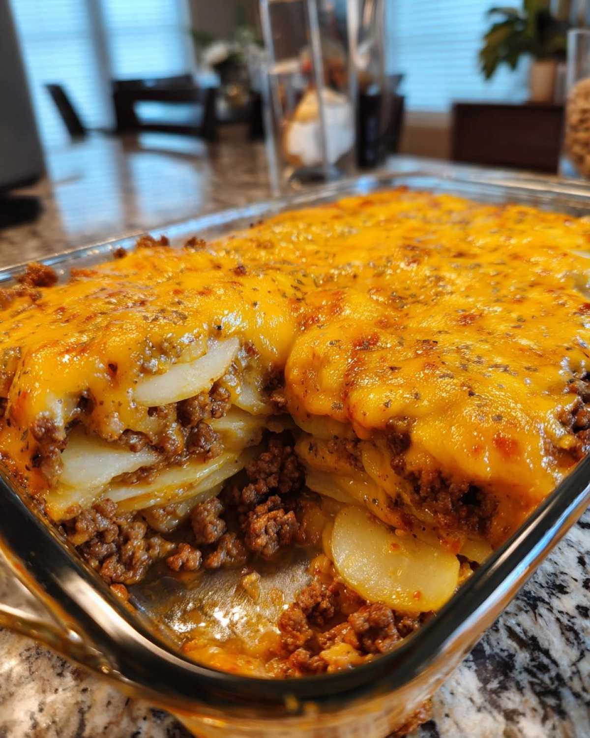 A close-up of a freshly baked hamburger potato casserole in a glass dish, featuring layers of sliced potatoes and seasoned ground beef topped with melted cheddar cheese.