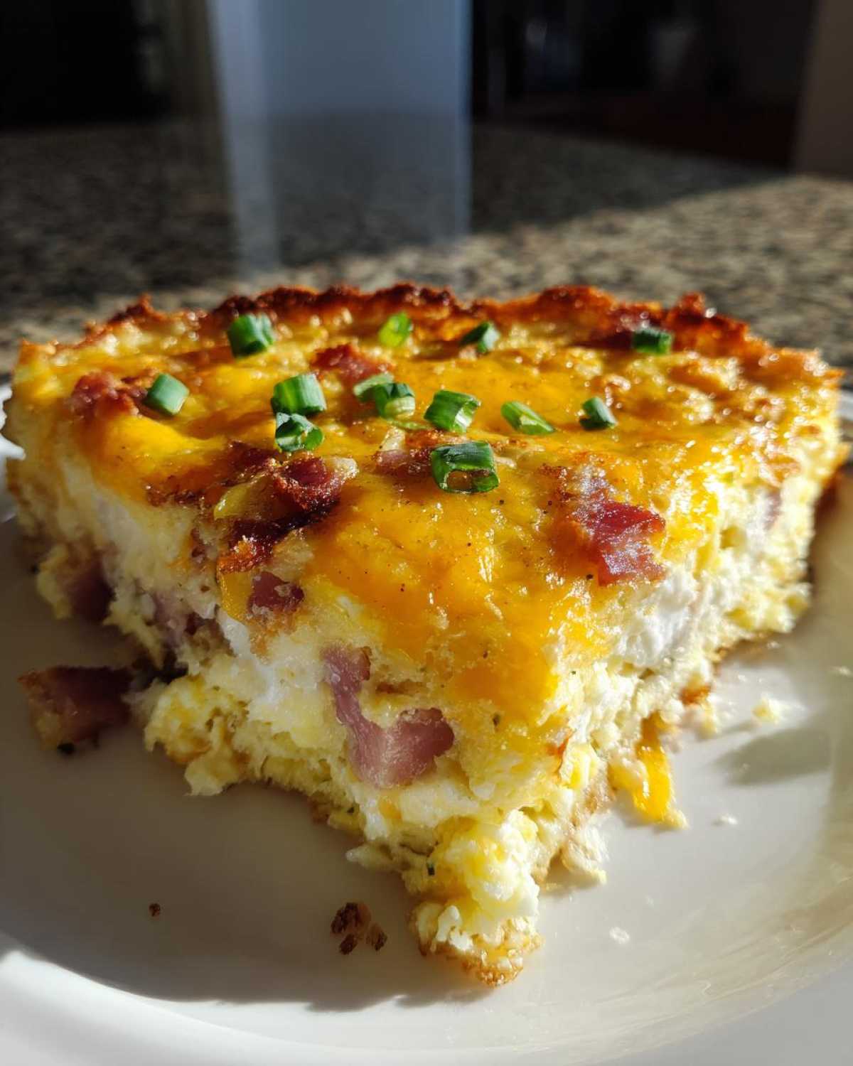 A slice of hash brown egg casserole topped with melted cheese, bacon, and green onions.