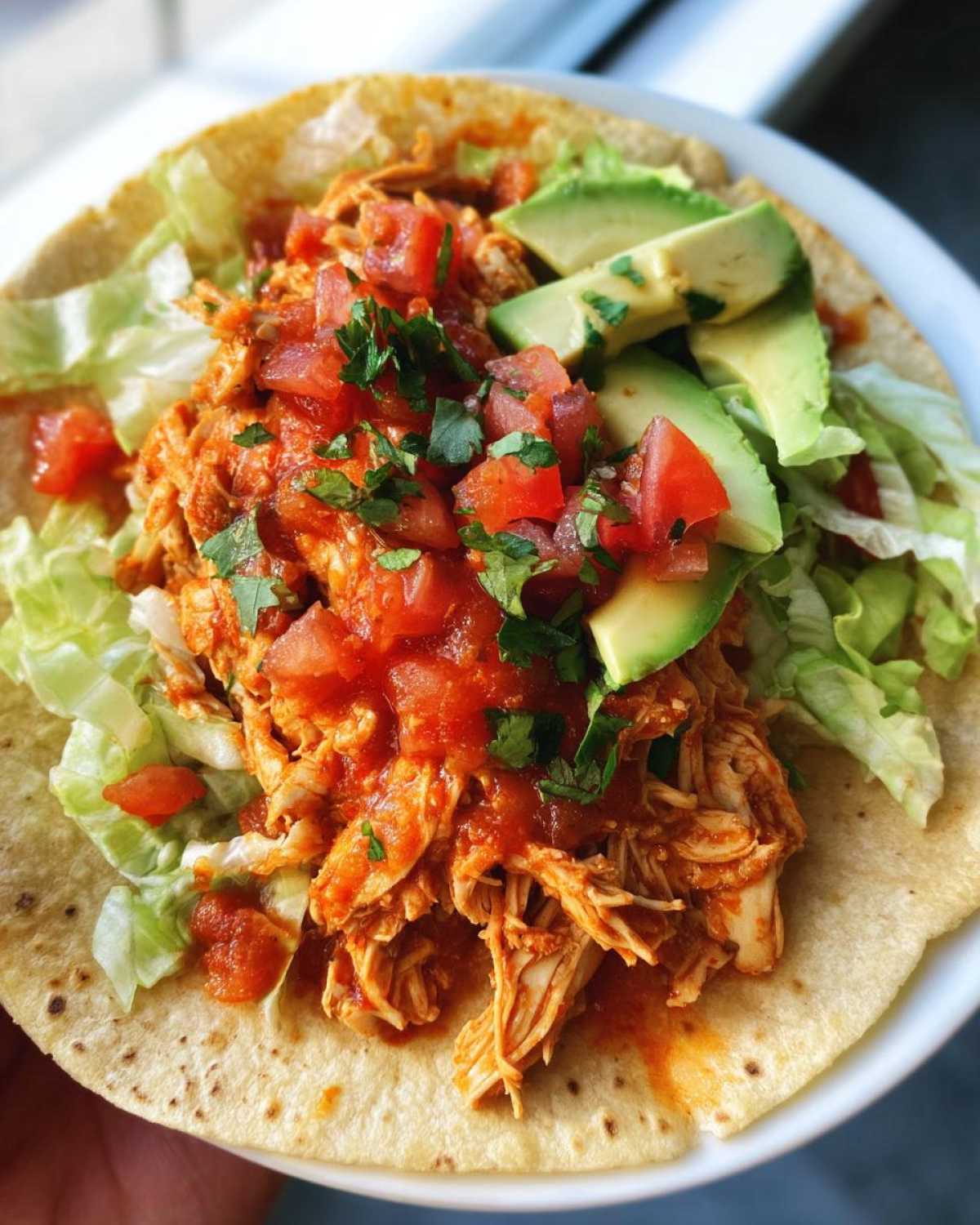 A delicious Healthy Crockpot Chicken Tacos served on a tortilla with shredded lettuce, diced tomatoes, avocado slices, and cilantro.